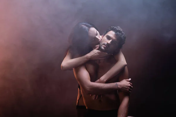 Mujer sensual abrazando y besando joven hombre sin camisa mirando a la cámara en el fondo oscuro con humo - foto de stock