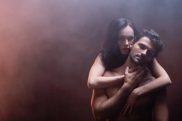 Seductive woman looking at camera while hugging shirtless man on dark background with smoke — Stock Photo