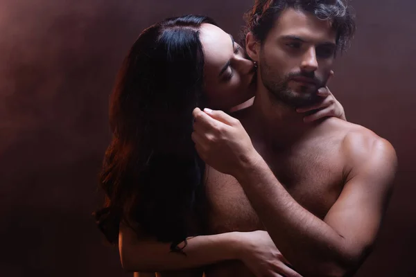 Sexy brünette Frau umarmt und küsst hemdlosen Mann auf dunklem Hintergrund — Stockfoto