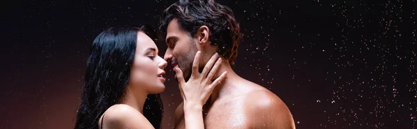 Vue latérale de femme sexy près de jeune homme torse nu sous la pluie sur fond sombre, bannière — Photo de stock