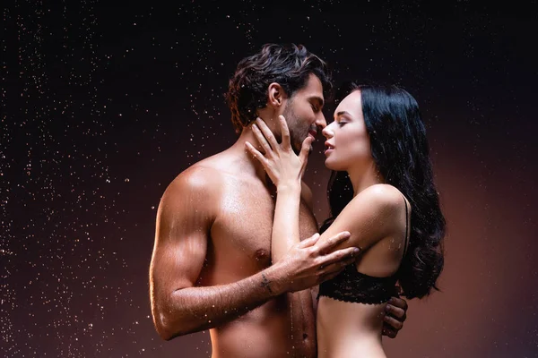 Jeune homme torse nu et femme brune sexy étreignant sous la pluie sur fond sombre — Photo de stock