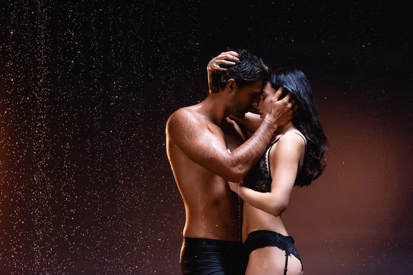 Vista laterale di uomo senza maglietta e donna bruna in biancheria intima nera che abbraccia e bacia sotto la pioggia su sfondo scuro — Foto stock