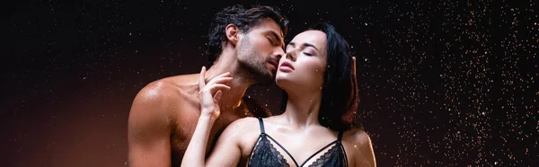 Young shirtless man kissing seductive woman in black lace bra on dark background under rain, banner — Stock Photo