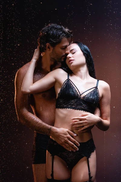 Hombre joven abrazando a mujer seductora en lencería de encaje negro bajo la lluvia sobre fondo oscuro - foto de stock