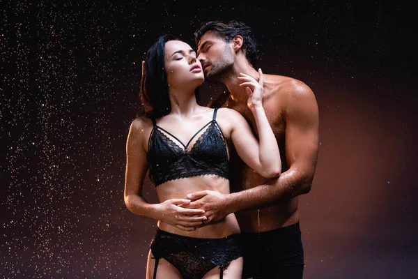 Hombre sin camisa abrazando y besando a mujer sexy en ropa interior negra bajo lluvia cayendo sobre fondo oscuro - foto de stock