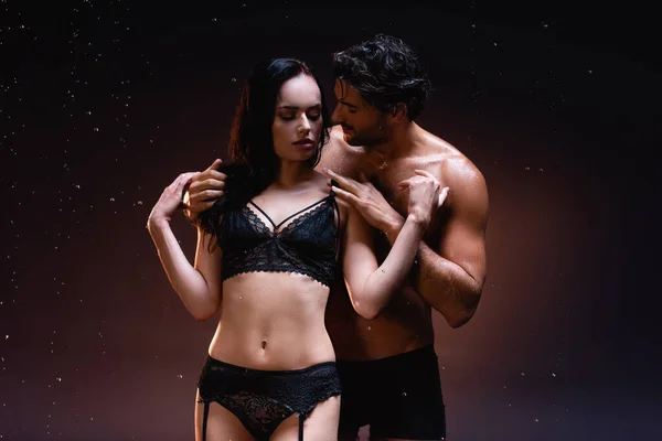 Sexy shirtless man touching sensual woman in black lace underwear on dark background under falling raindrops — Stock Photo