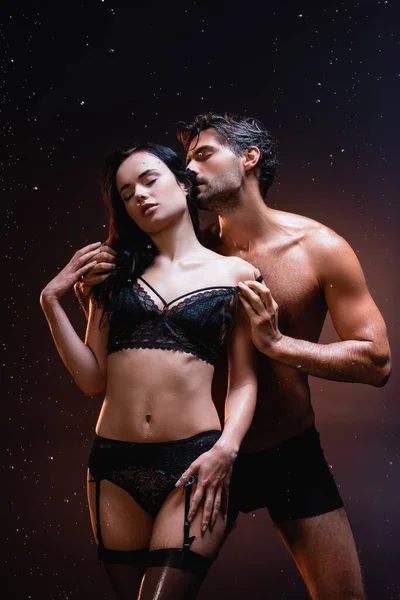 Jeune homme torse nu touchant et embrassant femme sexy en lingerie noire et bas sous la pluie sur fond sombre — Photo de stock