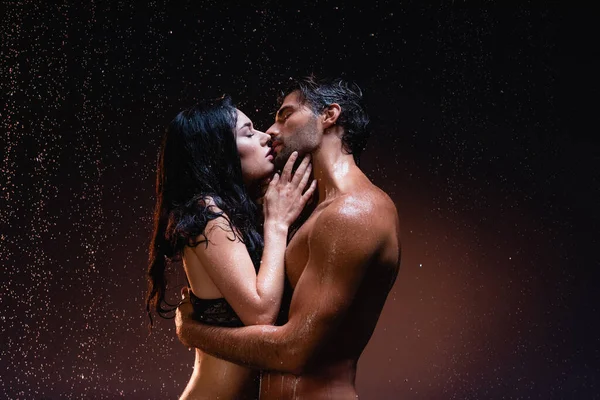 Side view of sexy shirtless man and brunette woman hugging and kissing on dark background under falling rain — Stock Photo