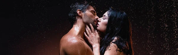 Vista lateral de mujer morena sexy y hombre sin camisa besándose con los ojos cerrados sobre fondo oscuro bajo gotas de lluvia, bandera - foto de stock