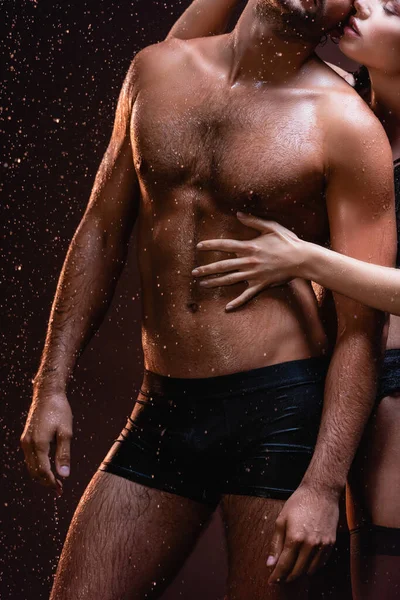 Ausgeschnittene Ansicht einer Frau, die den Oberkörper eines schulterfreien, muskulösen Mannes unter Regen auf dunklem Hintergrund berührt — Stockfoto