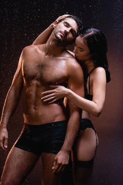 Sensual mujer abrazando sexy, musculoso hombre bajo lluvia cayendo sobre oscuro fondo - foto de stock