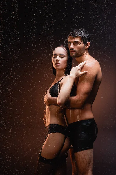 Shirtless muscular homem olhando para a câmera enquanto abraçando sexy mulher sob chuva no escuro fundo — Fotografia de Stock