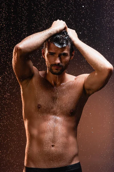 Sem camisa, homem muscular olhando para a câmera sob queda de gotas de chuva no fundo escuro — Fotografia de Stock