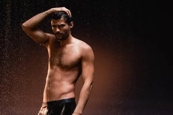 Sexy sin camisa hombre en negro calzoncillos posando bajo la lluvia sobre oscuro fondo - foto de stock