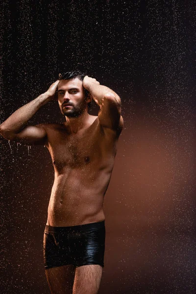 Hombre musculoso sin camisa en calzoncillos negros tocando la cabeza bajo la lluvia sobre fondo oscuro - foto de stock