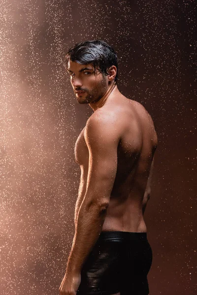 Сексуальный мужчина с мускулистым туловищем смотрит в камеру, позируя под дождем на тёмном фоне — стоковое фото