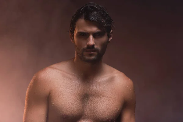 Mojado, hombre sin camisa mirando a la cámara mientras posando sobre fondo oscuro con humo - foto de stock