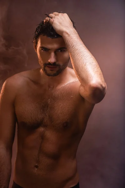 Sexy, hombre mojado con torso muscular posando con la mano en la cabeza sobre fondo oscuro con humo - foto de stock