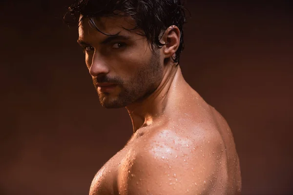 Wet, sexy man looking at camera while posing on dark background — Stock Photo
