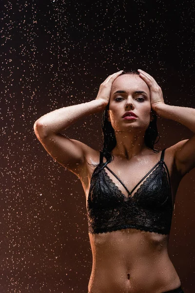Giovane donna seducente in lingerie pizzo nero toccare la testa sotto la pioggia su sfondo scuro — Foto stock