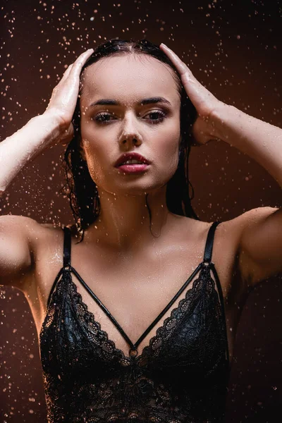 Mulher sensual em sutiã de renda preta tocando o cabelo enquanto olha para a câmera sob a chuva no fundo escuro — Fotografia de Stock