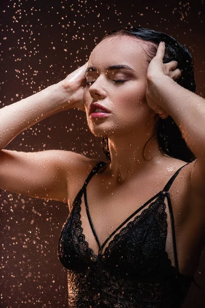 Соблазнительная женщина в черном кружевном бюстгальтере позирует с закрытыми глазами под проливным дождем на темном фоне — стоковое фото