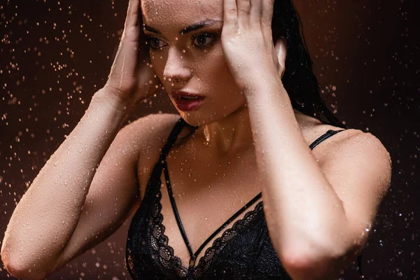 Sensual woman in black lace bra posing with hands near face under rain on dark background — Stock Photo