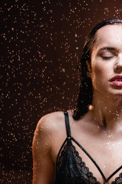 Чувственная женщина в черном кружевном лифчике с закрытыми глазами под проливным дождем на темном фоне — стоковое фото