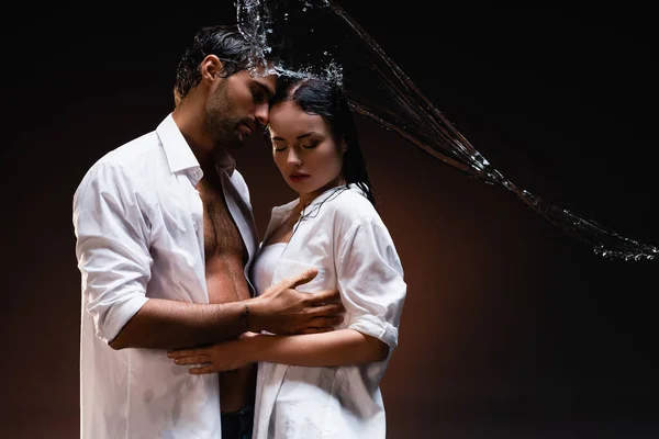 Young couple in white wet shirts embracing with closed eyes near water splashes on dark background — Stock Photo