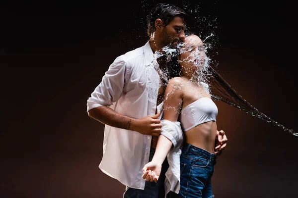 Homem de camisa molhada branca despir mulher sedutora perto de respingos de água no fundo escuro — Fotografia de Stock