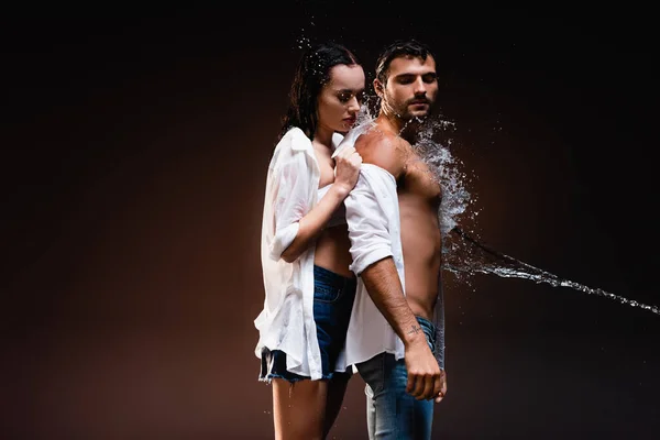 Woman in white wet shirt undressing muscular man near water splashes on dark background — Stock Photo