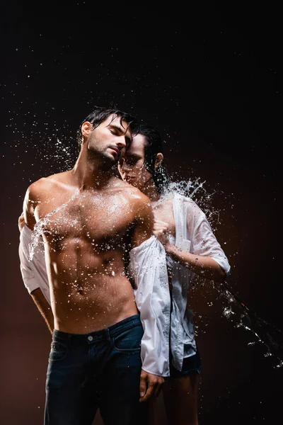 Mujer joven desnudándose sexy, hombre musculoso cerca de salpicaduras de agua sobre fondo oscuro - foto de stock