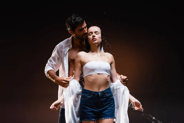 Young man taking off wet shirt from sexy girlfriend near water splashes on dark background — Stock Photo