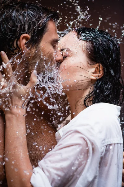 Joven hombre y mujer besándose con los ojos cerrados cerca de salpicaduras de agua sobre fondo oscuro - foto de stock