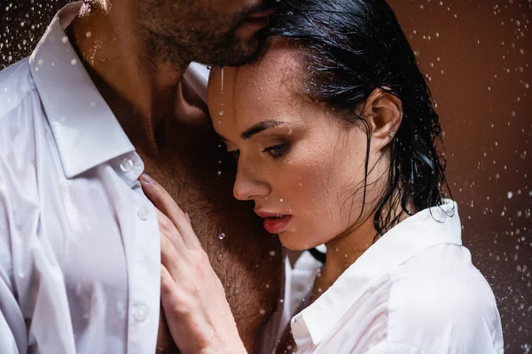 Mujer joven apoyada en el pecho del hombre mojado bajo la lluvia que cae sobre fondo oscuro - foto de stock