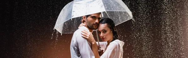 Joven pareja en camisas mojadas abrazando y mirando a la cámara mientras está de pie bajo la lluvia con paraguas sobre fondo oscuro, pancarta - foto de stock