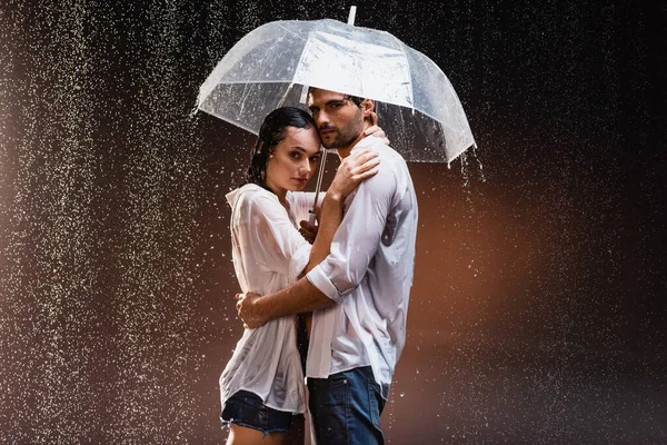 Pareja joven en camisas mojadas de pie bajo la lluvia con paraguas transparente sobre fondo oscuro - foto de stock