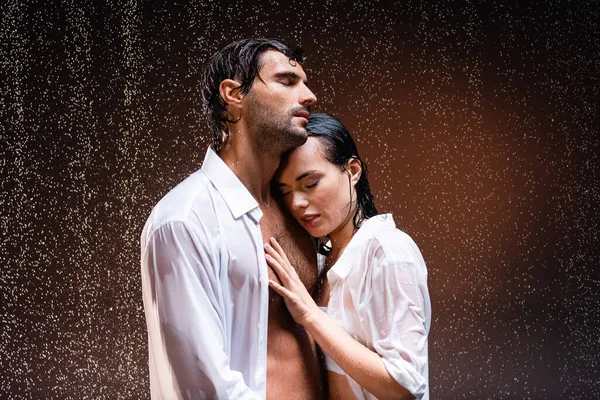 Jeune couple en chemises blanches debout sous la pluie avec les yeux fermés sur fond sombre — Photo de stock