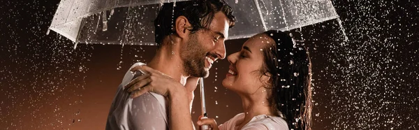 Side view of happy couple in wet shirts standing face to face under rain with transparent umbrella on dark background, banner — Stock Photo