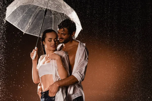 Sonriente hombre cogido de la mano en los bolsillos de la mujer sexy de pie bajo la lluvia con paraguas transparente sobre fondo oscuro - foto de stock