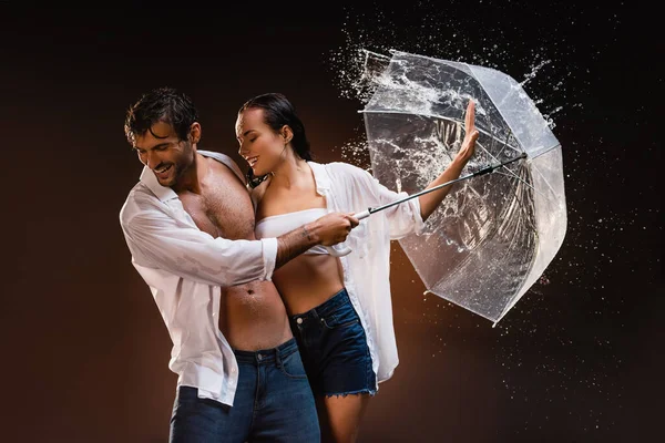 Cheerful couple in wet shirts, with transparent umbrella near water splashes on dark background — Stock Photo