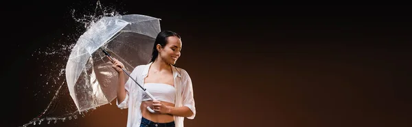 Щаслива, мокра жінка в білій сорочці, що стоїть з парасолькою біля водяних бризок на темному фоні, банер — стокове фото