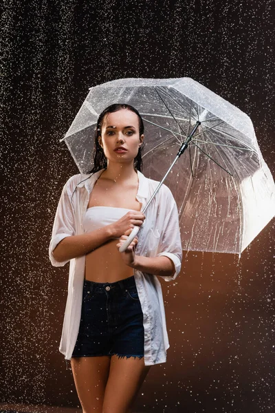 Sensual, wet woman looking at camera while standing with umbrella under rain on dark background — Stock Photo