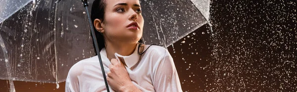 Wet woman looking away while standing with rain under transparent umbrella on dark background, banner — Stock Photo