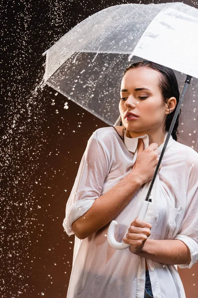 Mulher em camisa molhada de pé com guarda-chuva sob chuva no fundo escuro — Fotografia de Stock