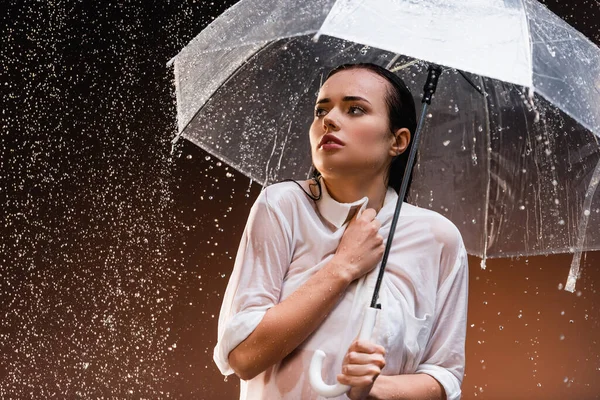 Mujer mojada mirando hacia otro lado mientras está de pie con paraguas transparente bajo la lluvia sobre fondo oscuro - foto de stock