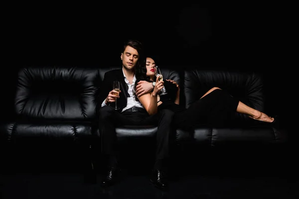 Sexy couple holding glasses of champagne while sitting on sofa isolated on black — Stock Photo