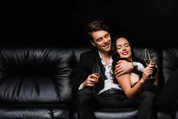 Усміхнена пара тримає келихи шампанського, відпочиваючи на дивані ізольовано на чорному — стокове фото