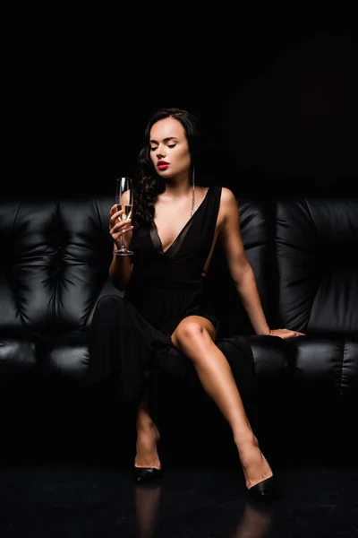 Seductive woman in dress sitting on sofa and holding glass of champagne on black — Stock Photo