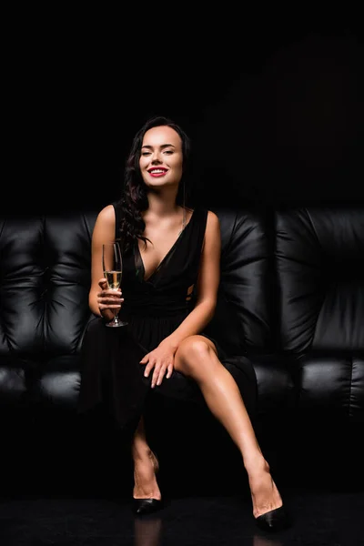 Cheerful woman in dress sitting on sofa and holding glass of champagne on black — Stock Photo